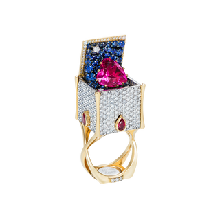 An ornate gold ring featuring a box-shaped pavé setting with a large heart-shaped pink gemstone on top, flanked by rows of blue and clear stones, with smaller pink gemstone accents on the sides, shown on a plain white background.
